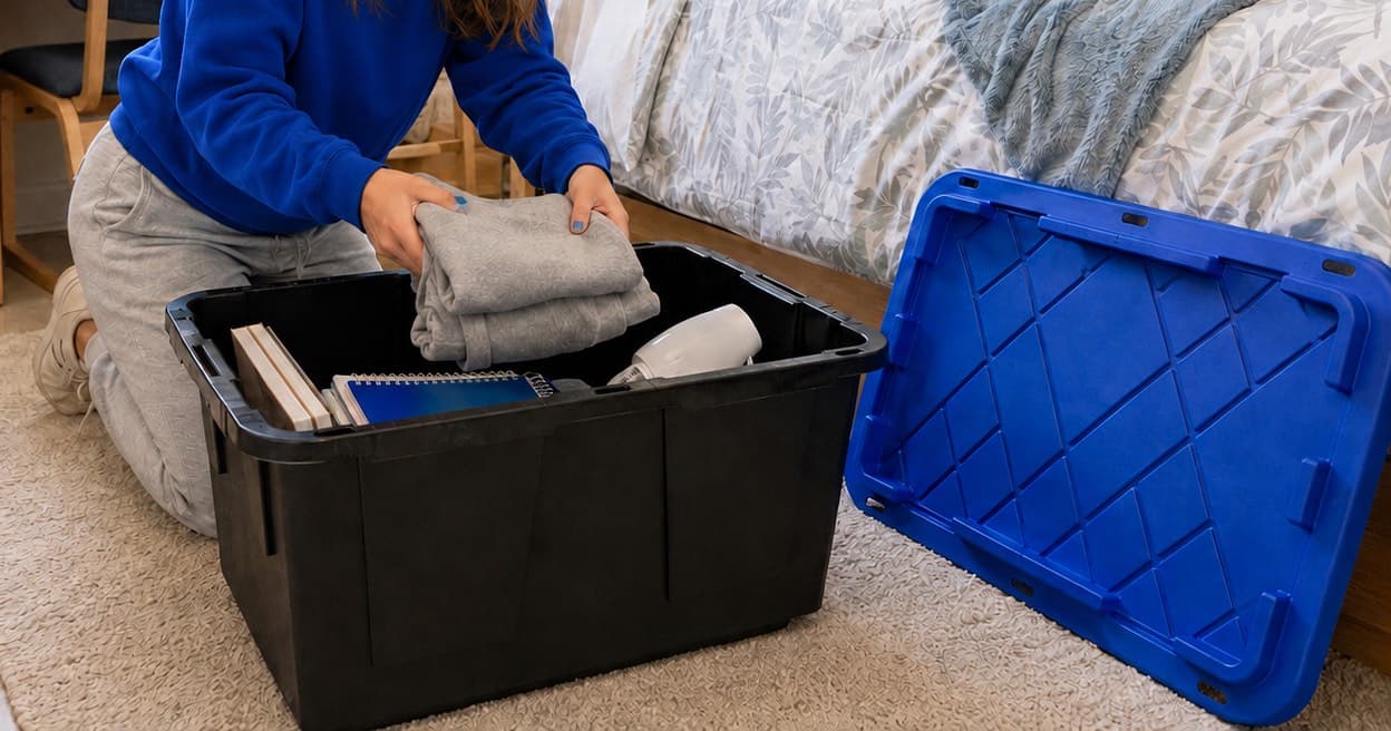 A student packing folded clothes and notebooks into a black moving tote in a dorm room, with the royal blue lid propped alongside